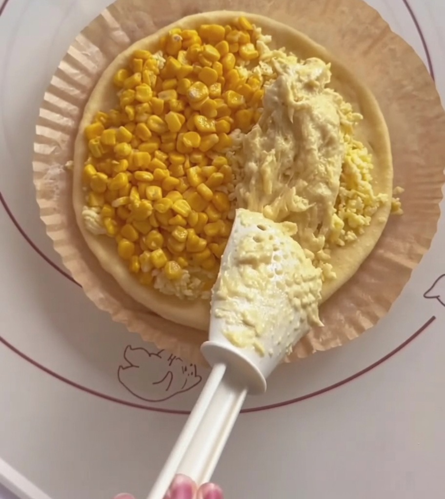 Une spatule blanche étalant de la pâte de durian jaune crémeuse sur une moitié de la base de la pizza.