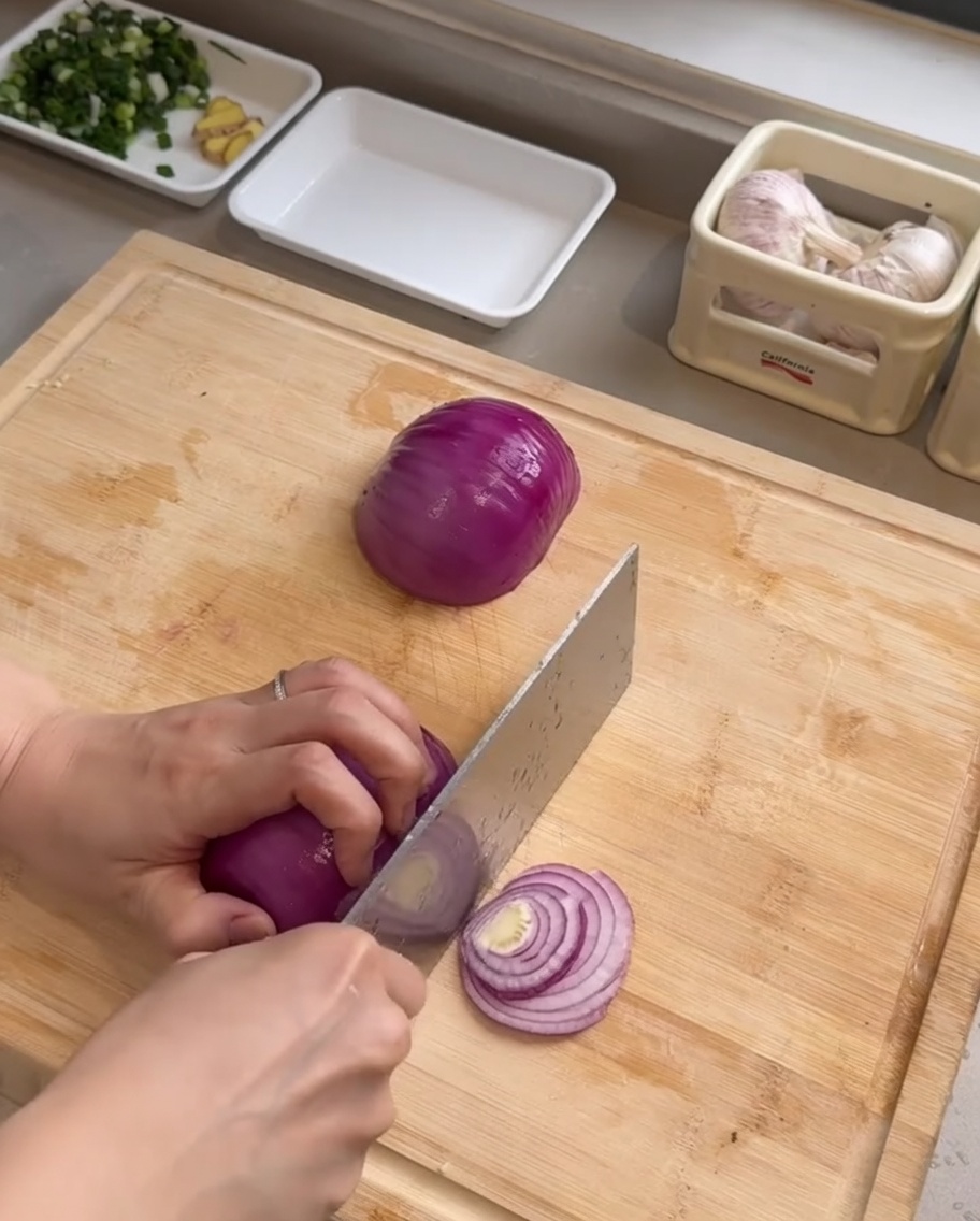 Un couteau de chef tranchant un oignon rouge en lamelles fines sur une planche en bois.