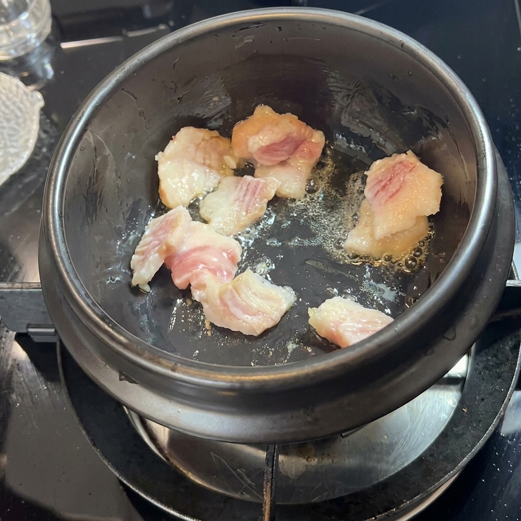Morceaux de poitrine de porc grésillant et rendant leur gras dans un pot en pierre noire.
