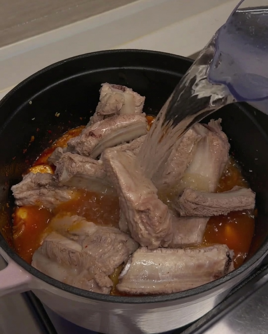 Eau claire versée depuis un pichet dans une marmite, recouvrant totalement les travers de porc et le kimchi.