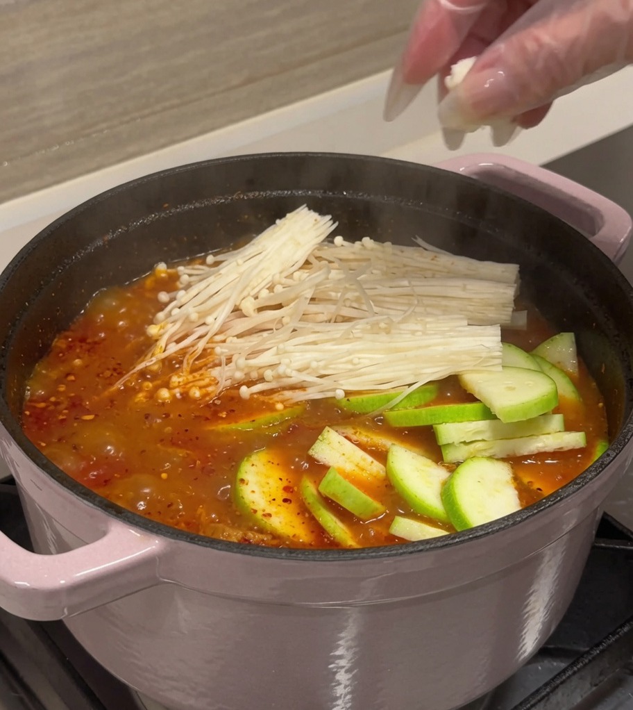 Tranches de courgettes, cubes de tofu et champignons enoki ajoutés dans une marmite de bouillon rouge bouillonnant.