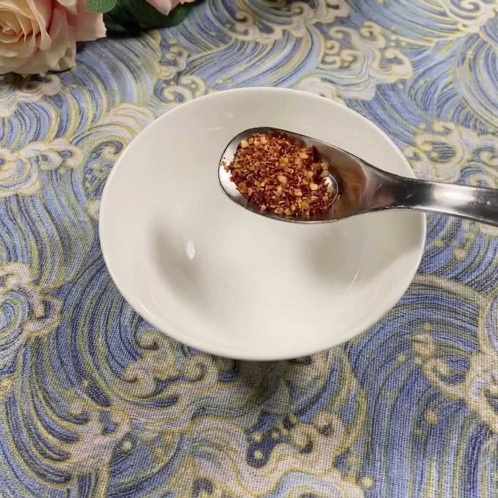 Une cuillère de poudre de piment grossière au-dessus dun bol blanc vide sur une surface bleue à motifs.
