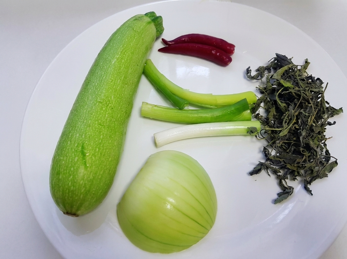 Ingrédients bruts dont une courge verte entière, des oignons verts, un oignon blanc, des feuilles de pérille et des piments rouges disposés sur une assiette blanche.