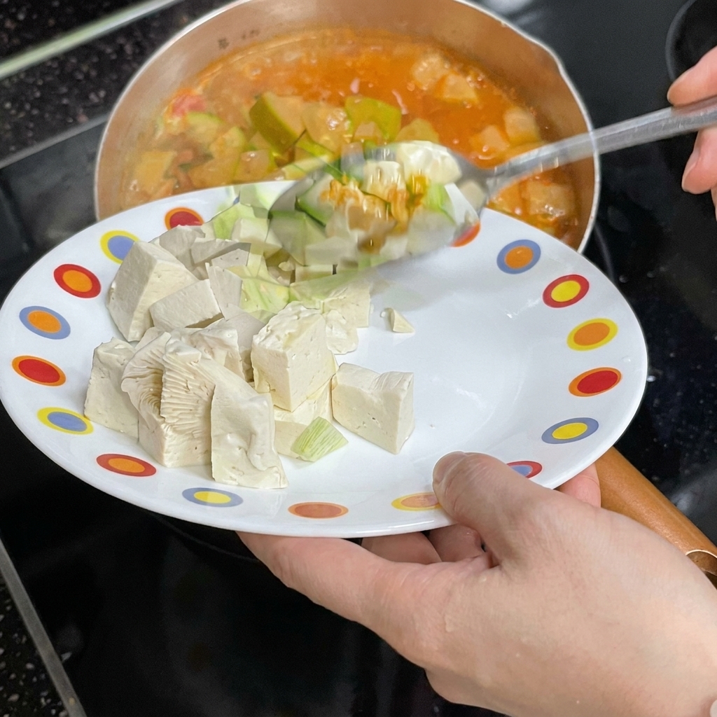 Une personne faisant glisser des cubes de tofu blanc depuis une assiette colorée dans le ragoût bouillonnant.