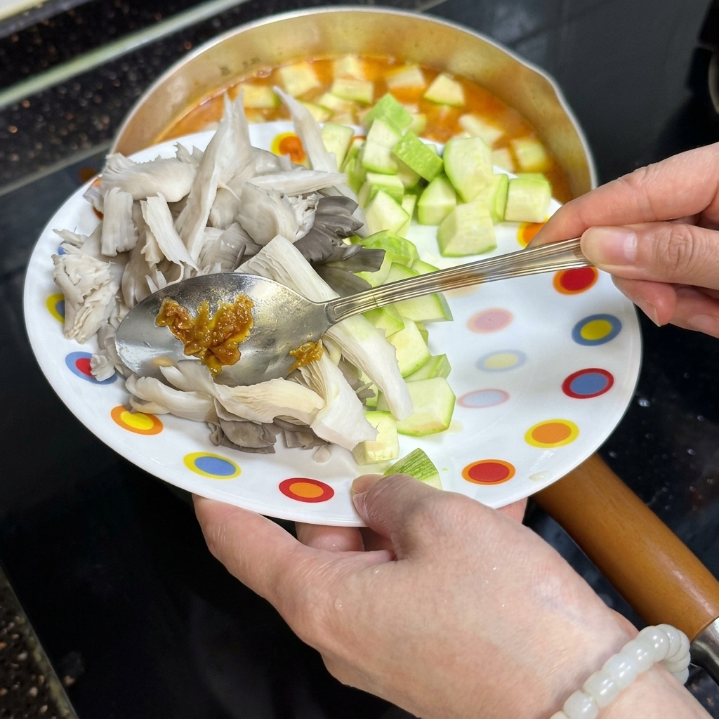 Une main utilisant une cuillère pour verser des dés de courgette et des pleurotes dans la marmite.