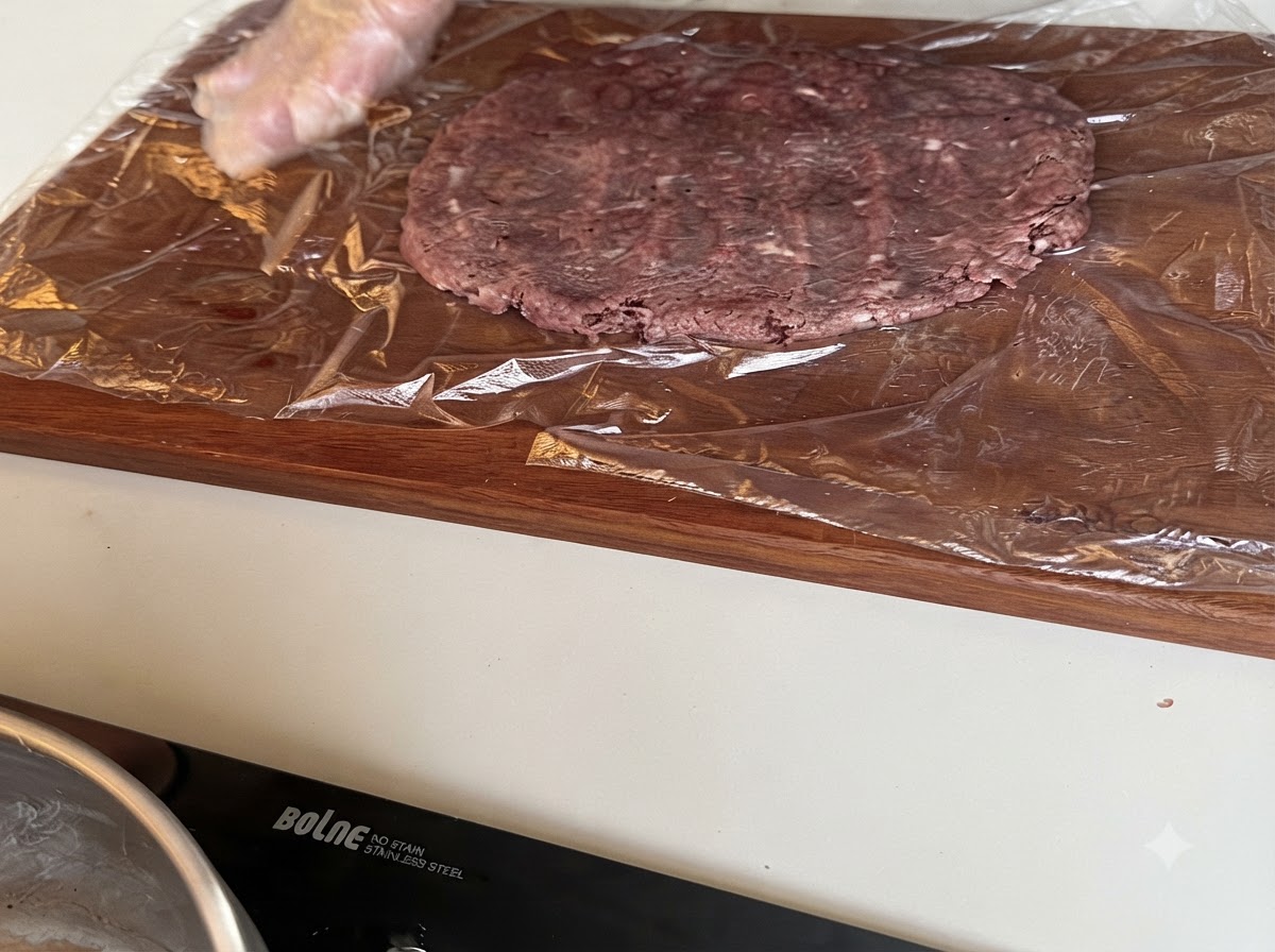 Un grand steak de bœuf cru en train dêtre aplati sur du film plastique étalé sur une planche à découper.
