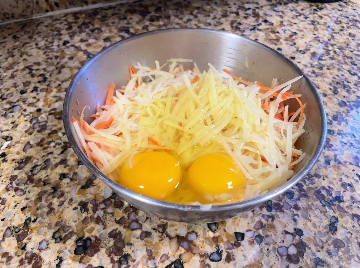Un bol en métal contenant des pommes de terre et des carottes fraîchement râpées, surmontées de deux œufs entiers.