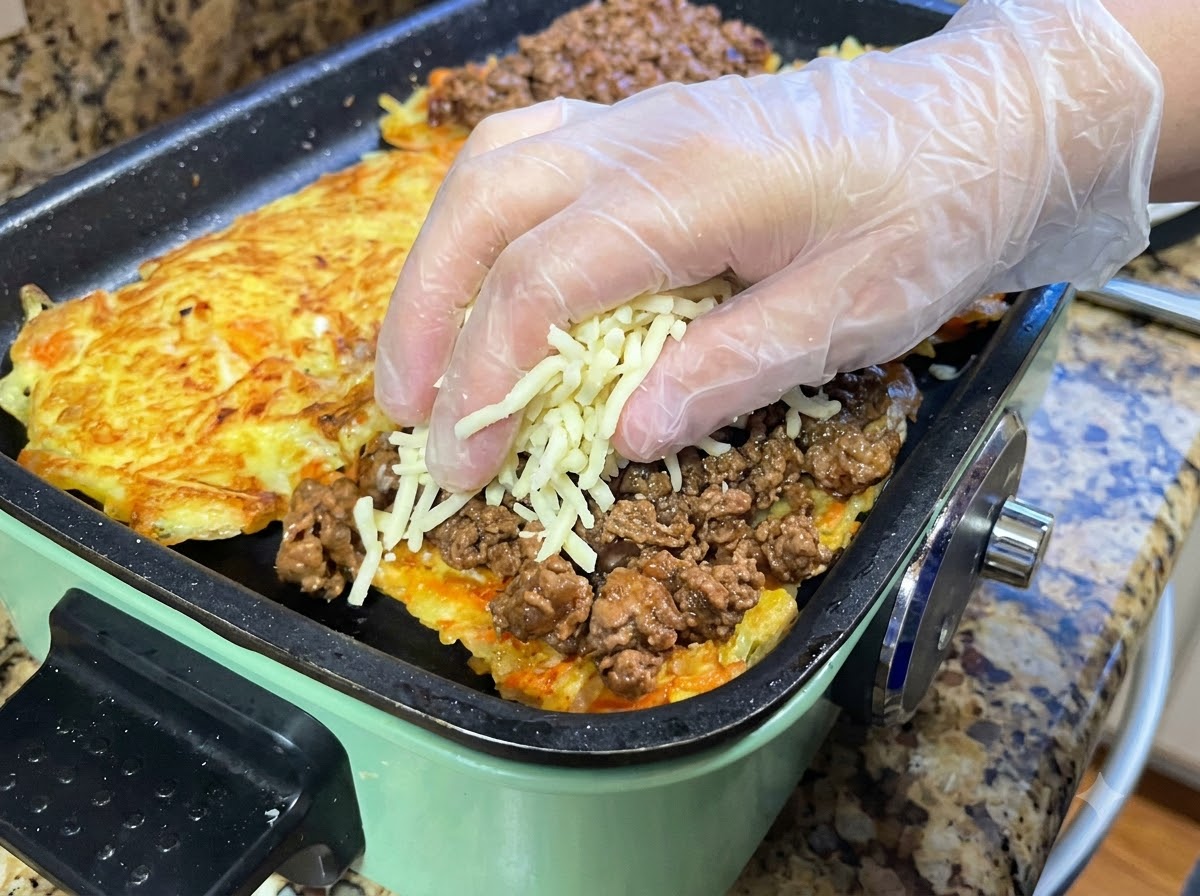 Des mains ajoutant du fromage râpé sur une couche de viande hachée recouvrant la galette de pommes de terre.