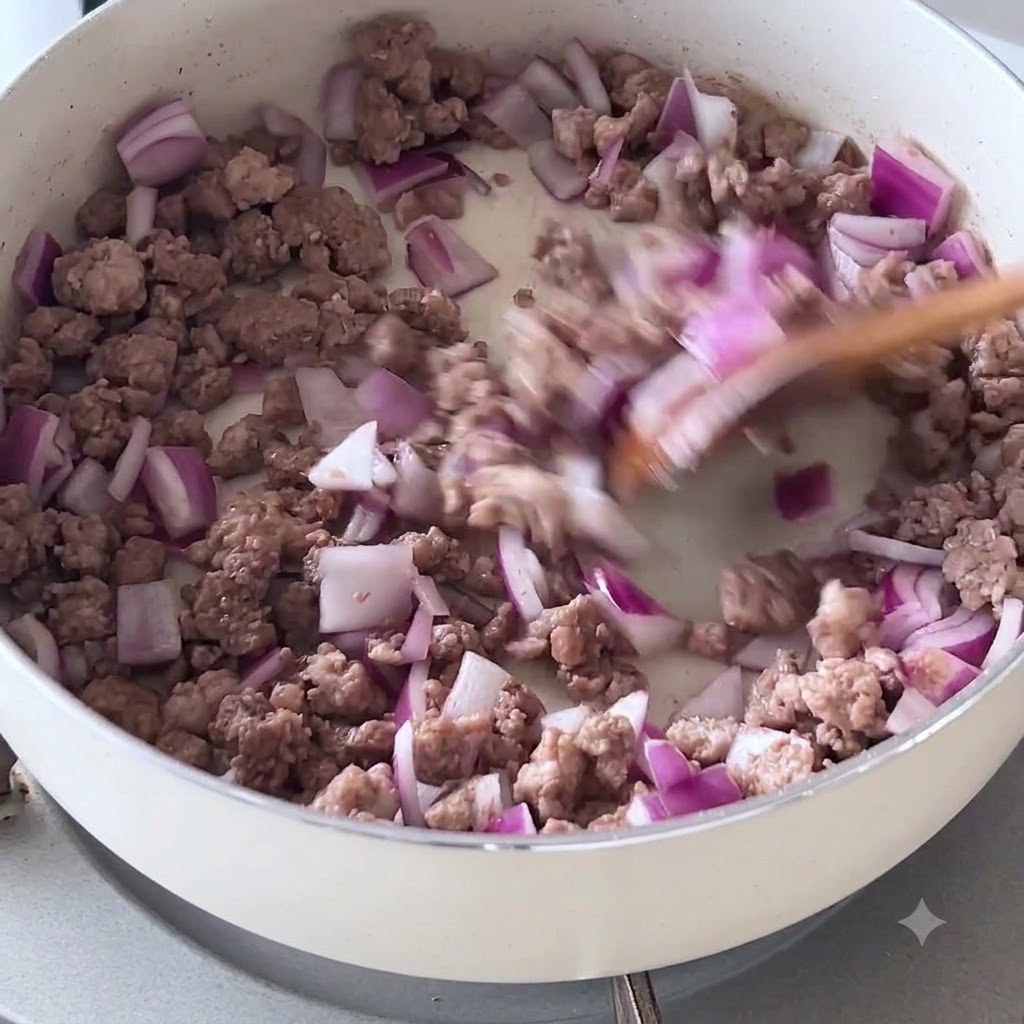 Oignons pourpres hachés et bœuf haché doré en train dêtre mélangés avec une spatule en bois dans une poêle blanche.