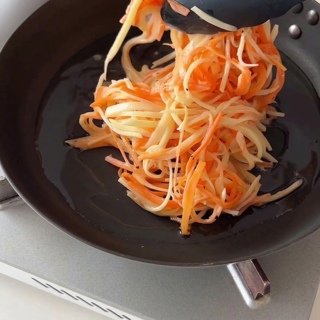Utilisation de pinces de cuisine noires pour placer un tas de pommes de terre et carottes râpées dans une poêle à frire huilée.