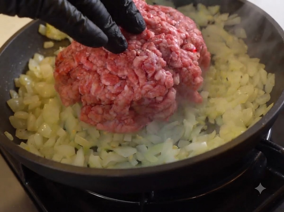 Une portion de bœuf haché cru placée sur des oignons sautés dans une poêle.