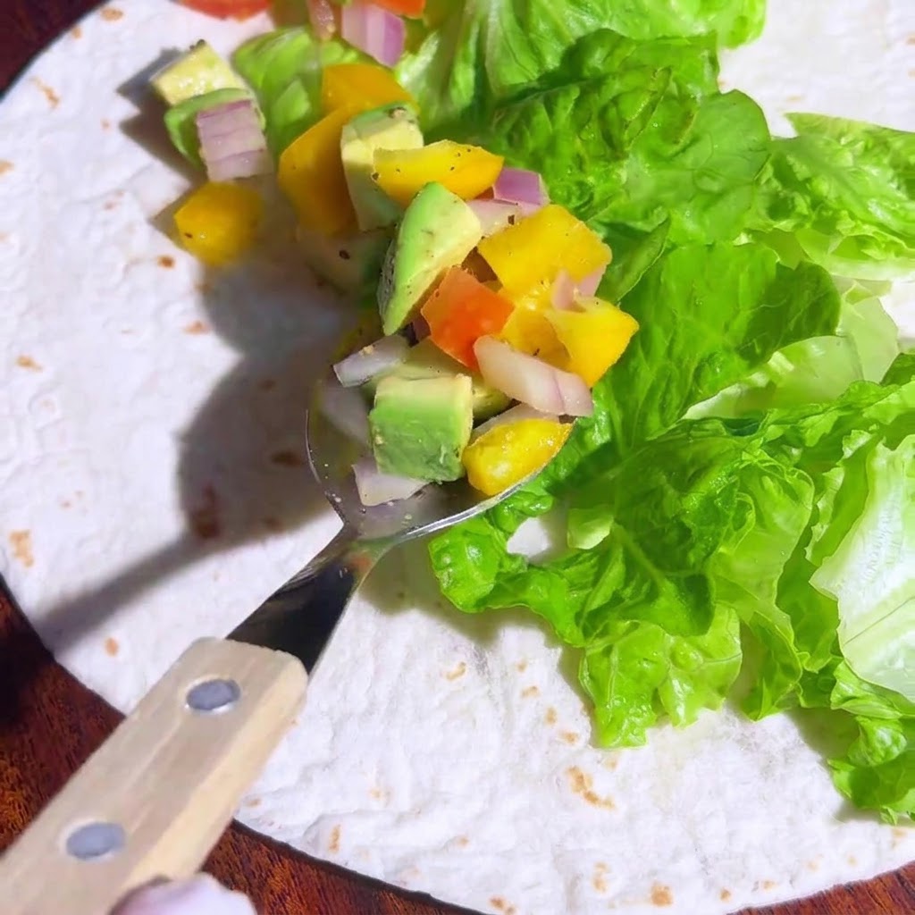 Une cuillère contenant de lavocat en dés coloré et une salsa de légumes, positionnée au-dessus de la laitue sur une tortilla.