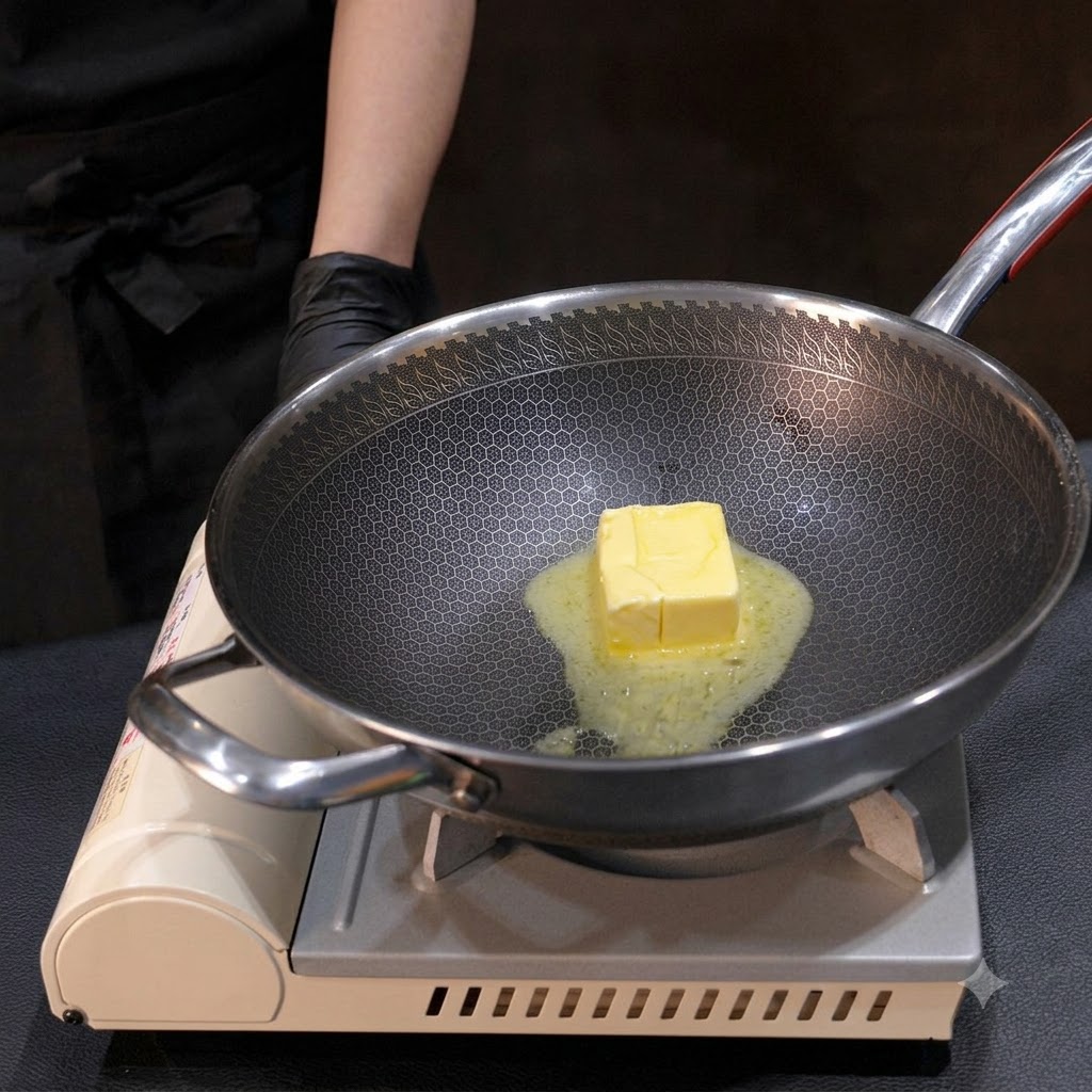 Un bloc de beurre jaune en train de fondre dans un wok en métal, remué avec un petit fouet sur le feu.