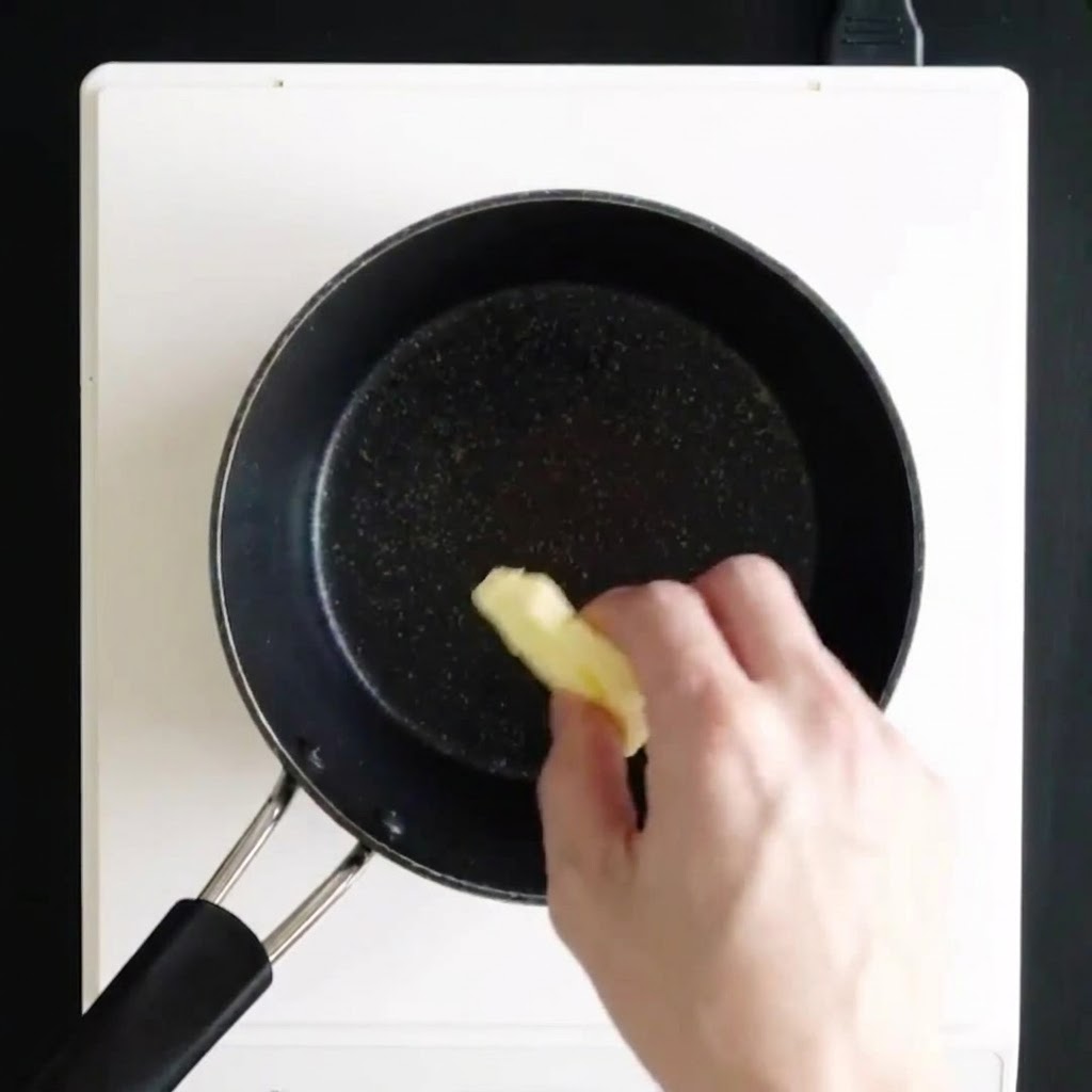 Une main déposant des morceaux de beurre dans une casserole noire sur une plaque de cuisson blanche pour les faire fondre.