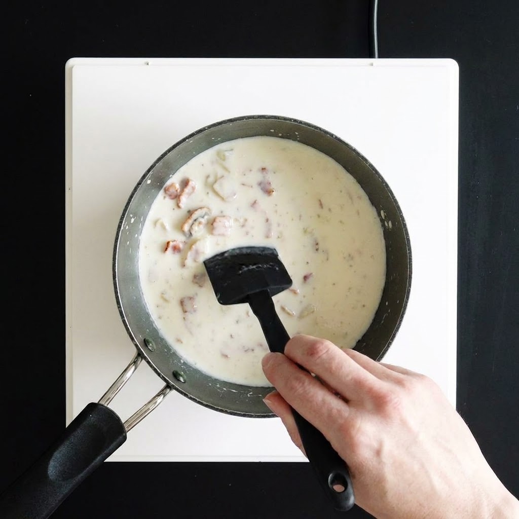 Une main remuant une soupe blanche épaisse et crémeuse dans une casserole à laide dune spatule en silicone noire.