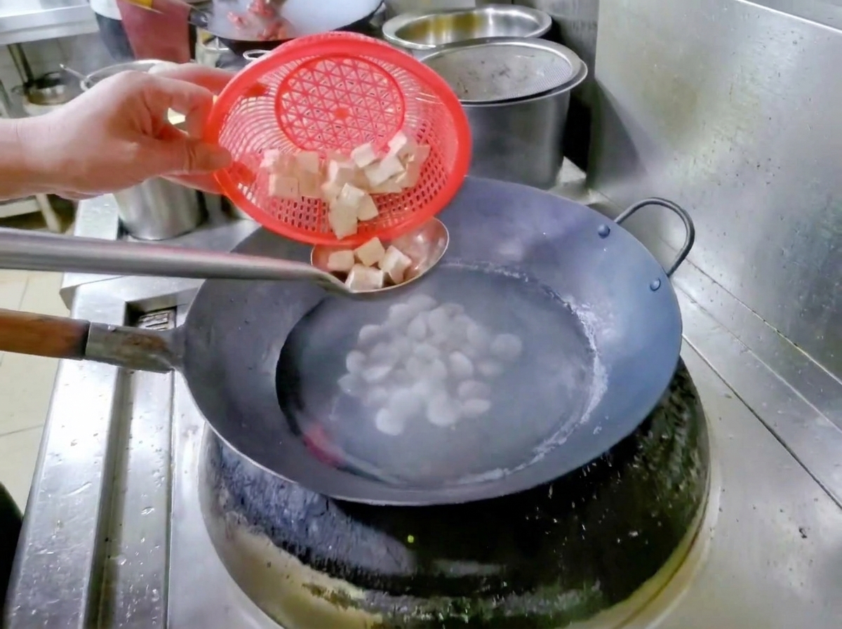 Une main tenant une passoire rouge, versant des dés de tofu dans un wok deau et de palourdes.