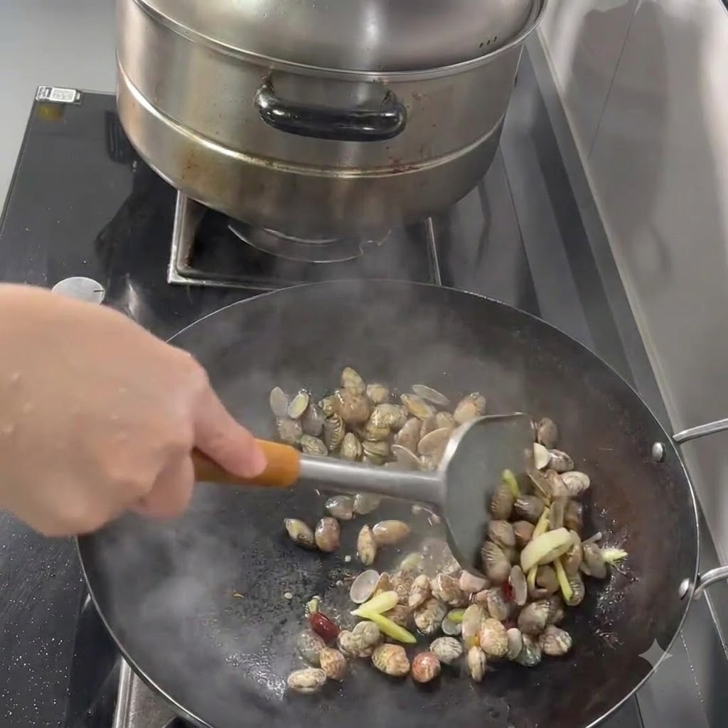 Palourdes, ail, gingembre et piments sautés dans un wok à laide dune spatule en métal.