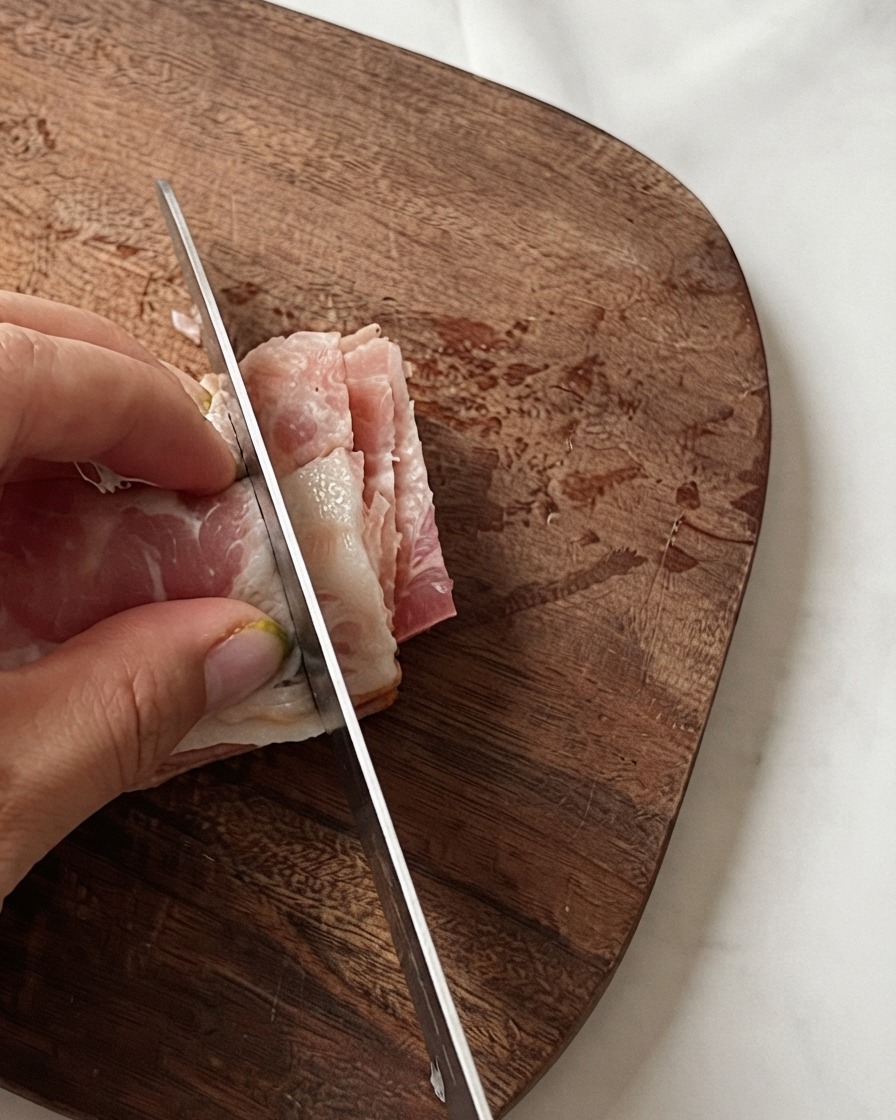 Les mains dun chef tenant des tranches de bacon empilées pendant quun couteau les coupe en petits morceaux sur une planche en bois.