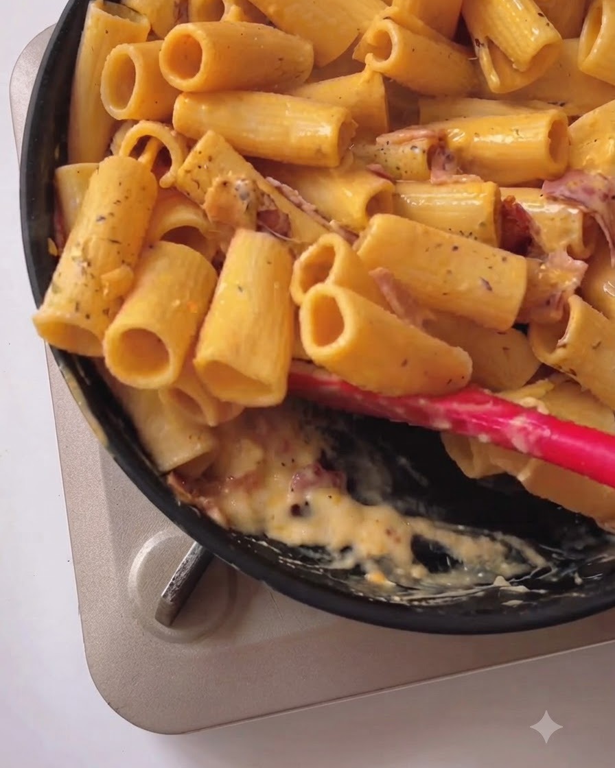 Des rigatoni mélangées avec une sauce jaune crémeuse et du bacon dans une poêle noire à laide dune spatule en silicone rouge.