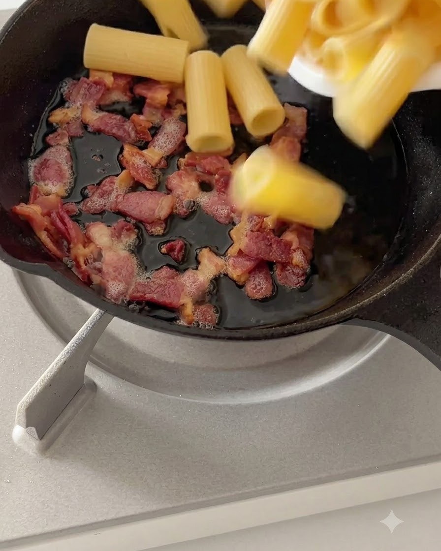 Des rigatoni bouillis mélangés avec du bacon cuit dans une poêle à feu doux.