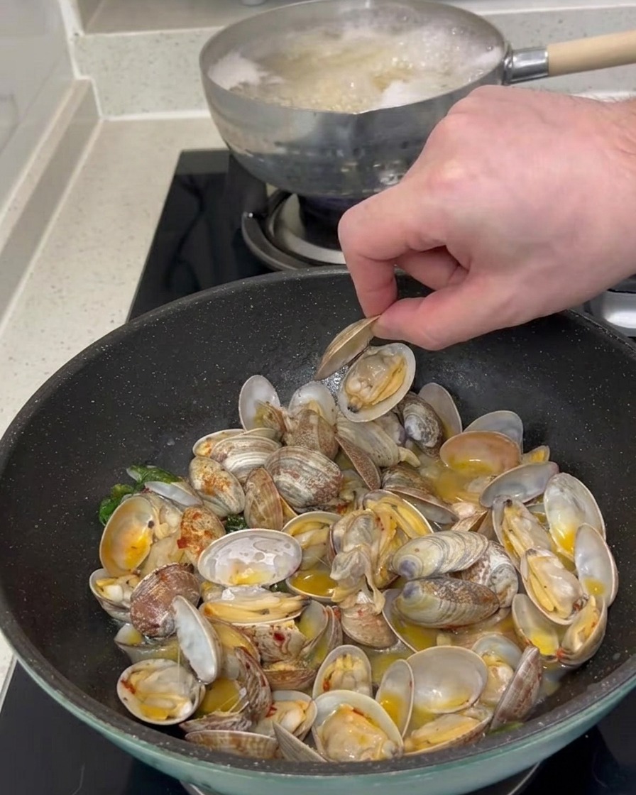 La main dune personne ramassant une coquille de palourde ouverte dans une poêle remplie de palourdes cuites et de sauce.