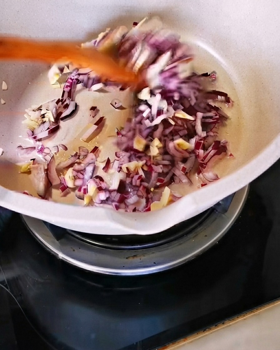 De loignon rouge haché et de lail émincé sautés avec une spatule en bois dans une poêle blanche.