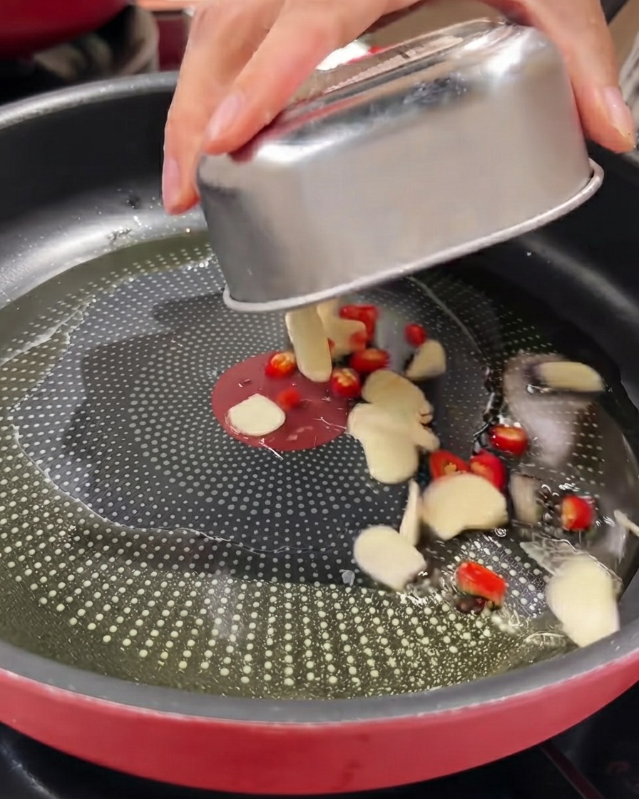 De lail tranché, des piments oiseaux rouges et de la ciboulette versés dun récipient en métal dans une poêle huilée.