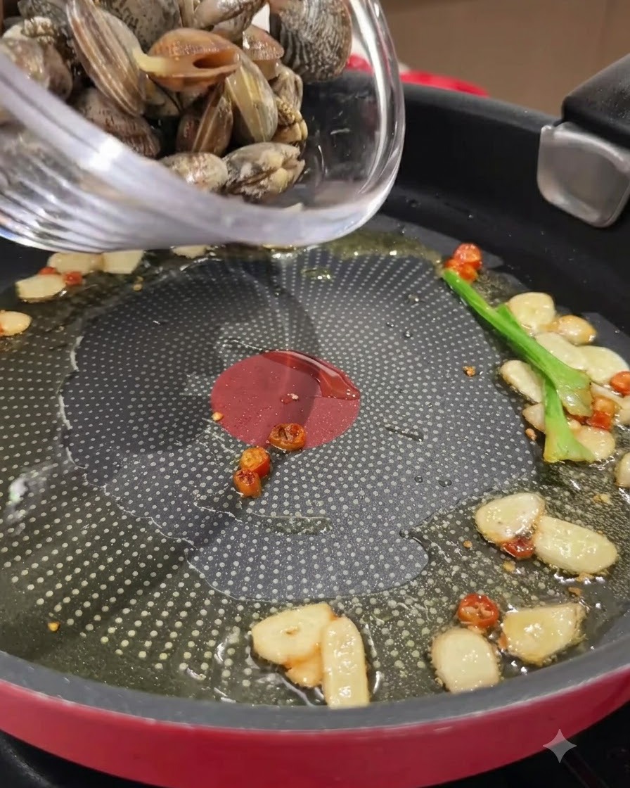 Des palourdes fraîches versées dun bol en verre dans une poêle contenant lail, le piment et lhuile sautés.