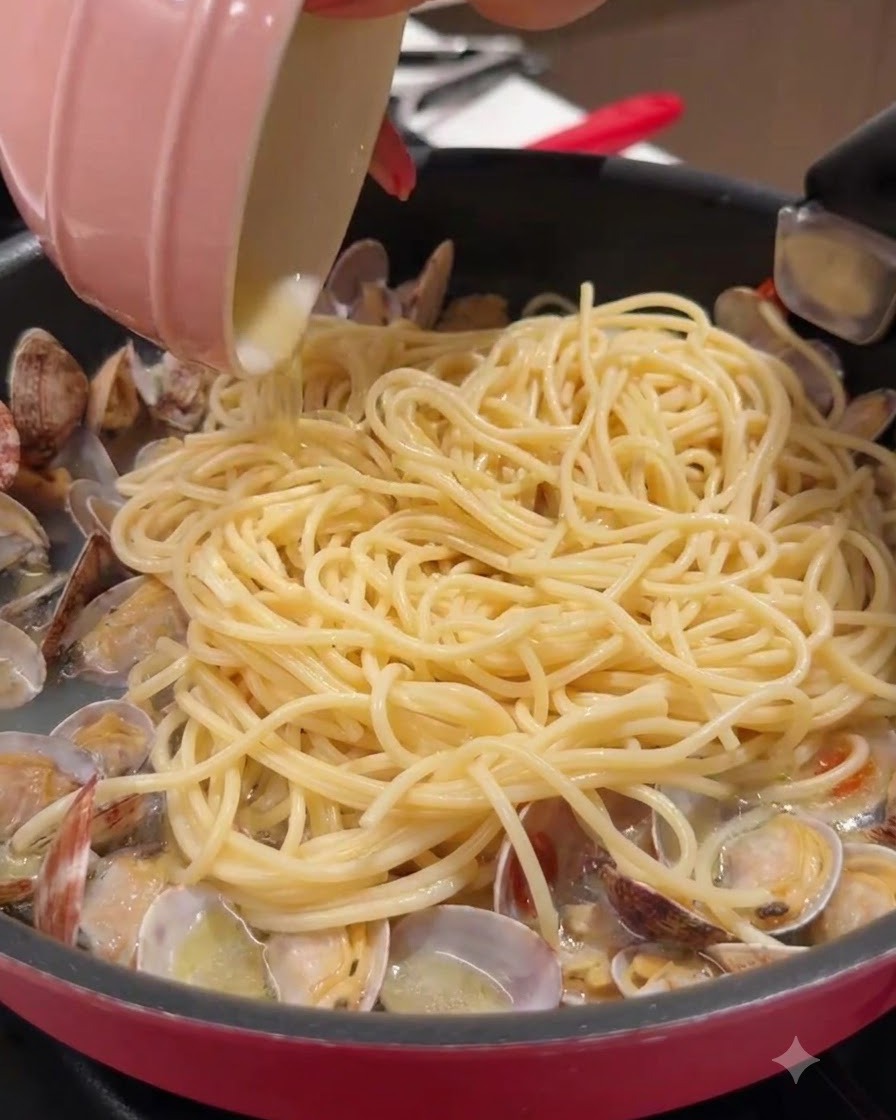Un petit bol rose versant du liquide dans une poêle remplie de spaghettis et de palourdes.