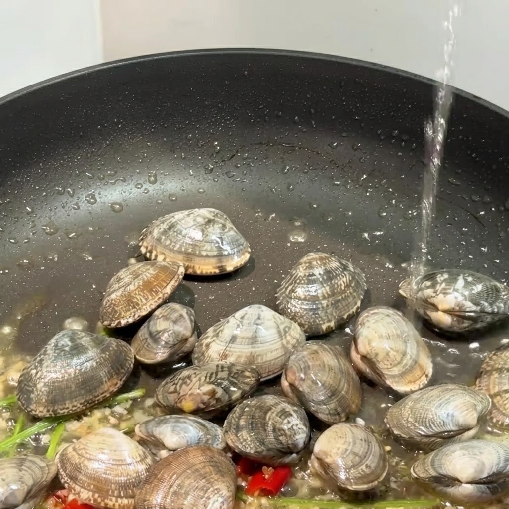 Une poêle noire contenant des palourdes, de lail et des piments, avec du liquide en train dy être versé.