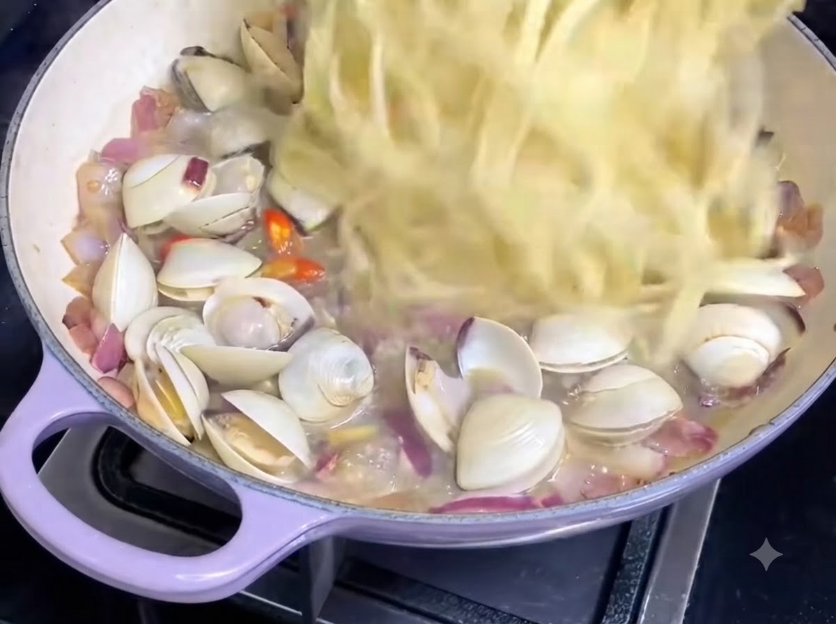 Brins de spaghetti cuits jetés dans une poêle contenant la sauce aux palourdes et les oignons.