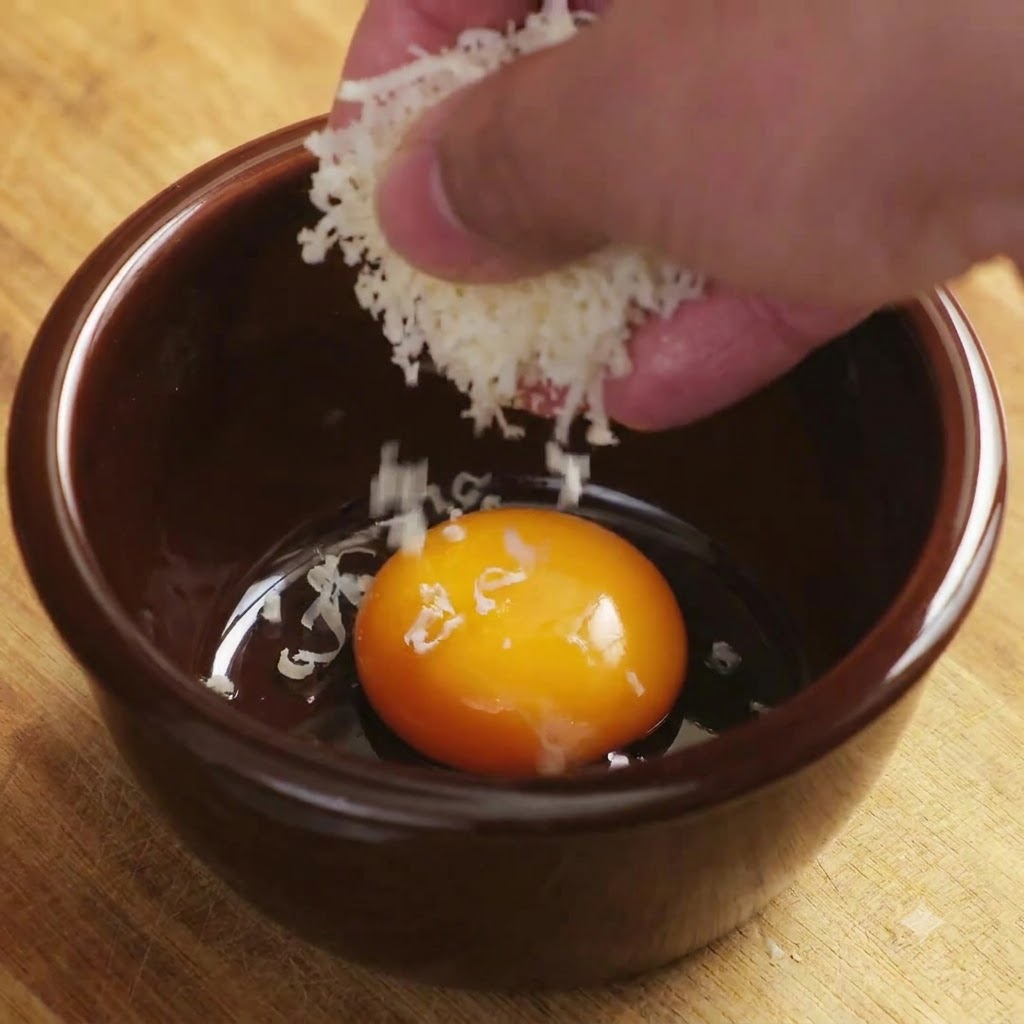Parmesan râpé tombant sur un jaune dœuf brillant dans un bol en céramique brune.