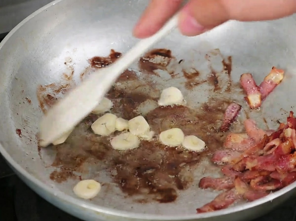 Ail émincé mélangé dans la poêle avec le bacon cuit à laide dune cuillère en bois.