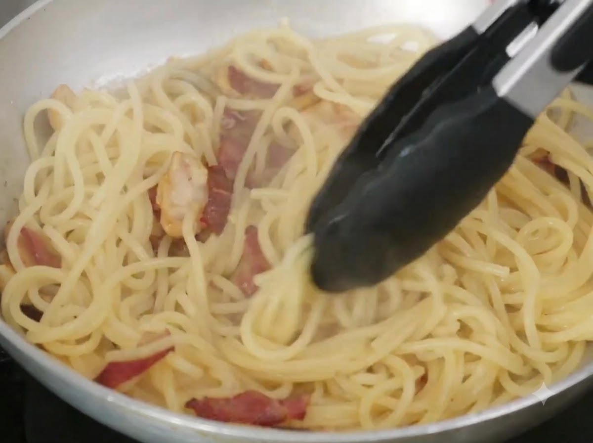 Beurre et eau de cuisson mélangés aux spaghettis dans une poêle à laide de pinces.