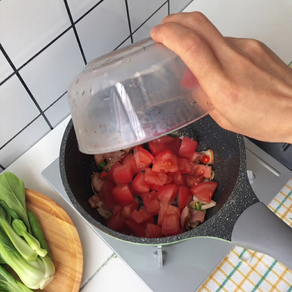 Main versant des morceaux de tomates dun bol en verre dans une poêle contenant du bacon frit et des oignons verts.