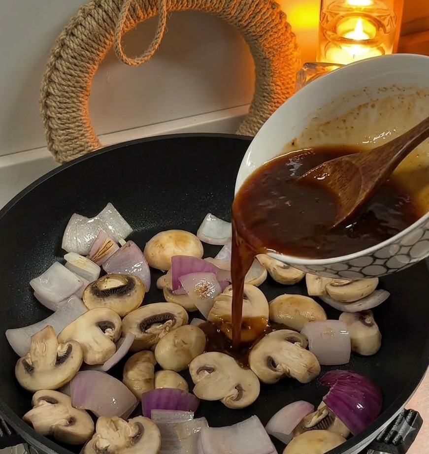 Versement dune sauce brune dun bol vers une poêle contenant des oignons tranchés et des champignons de Paris.