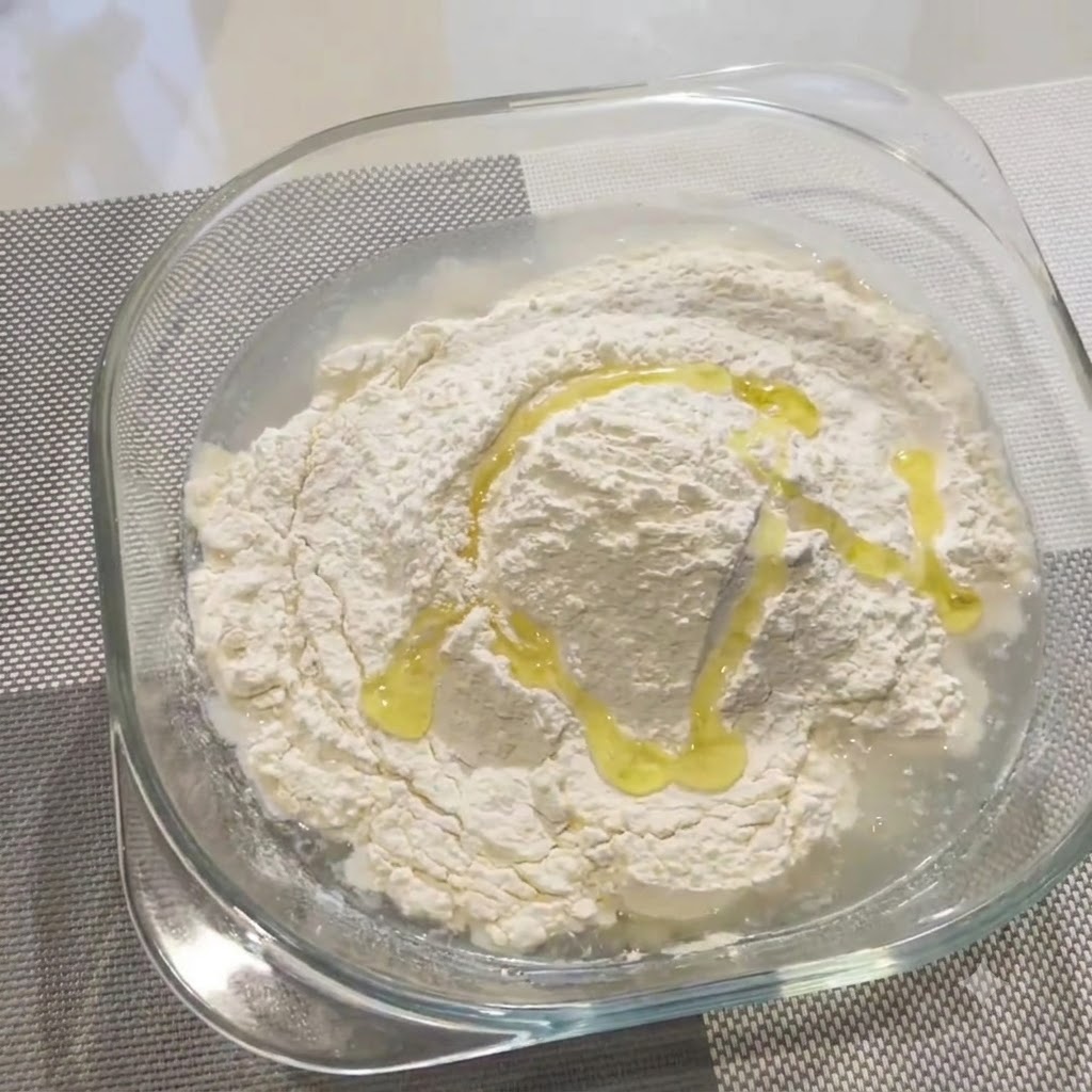Un filet dhuile dolive versé sur le mélange de farine et deau dans un bol en verre.