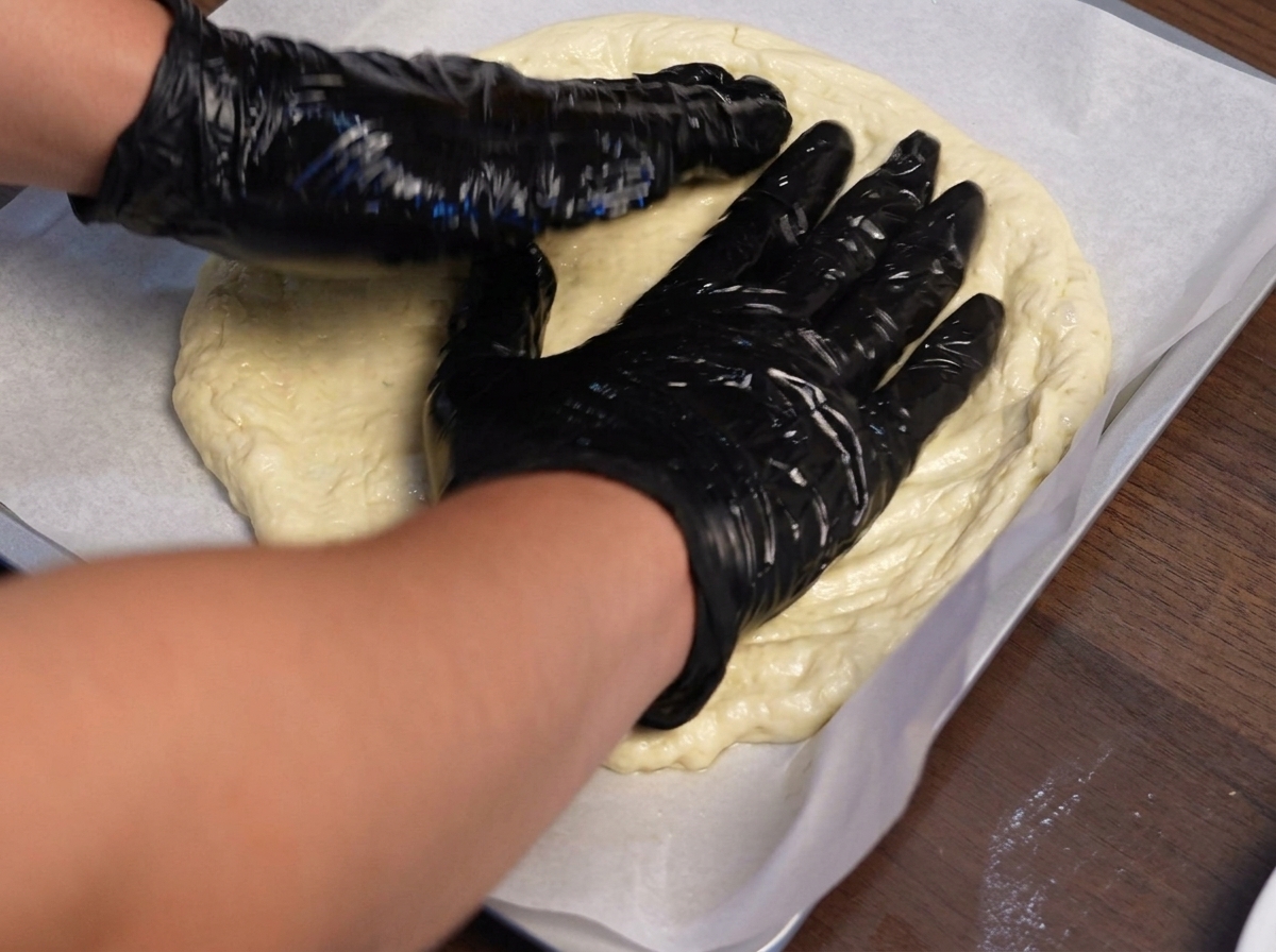 Mains gantées façonnant la pâte à pizza vers lextérieur sur une plaque de cuisson recouverte de papier sulfurisé.