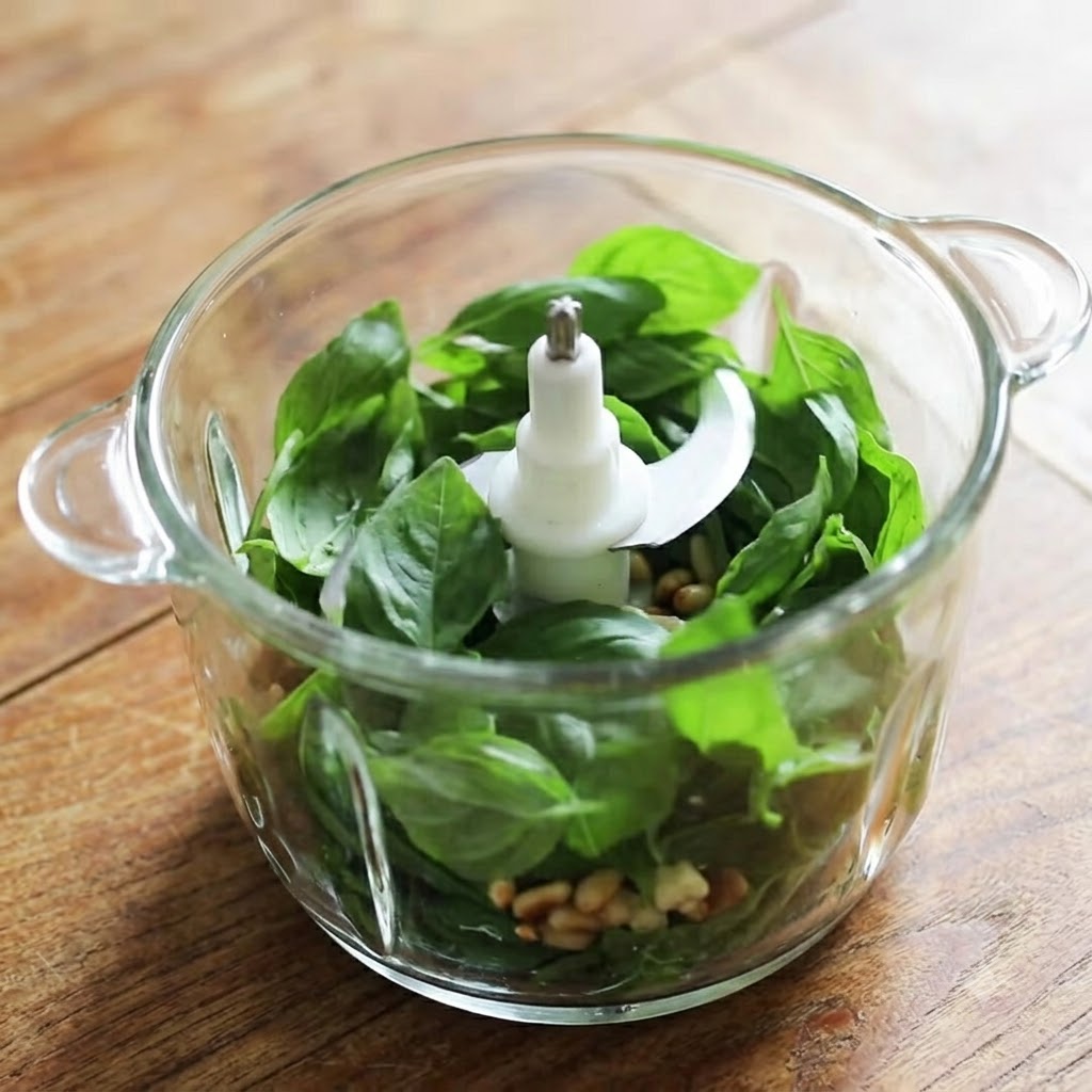 Un bol de mixeur en verre rempli de feuilles de basilic frais, de pignons de pin, dail et de fromage prêts à être mixés.