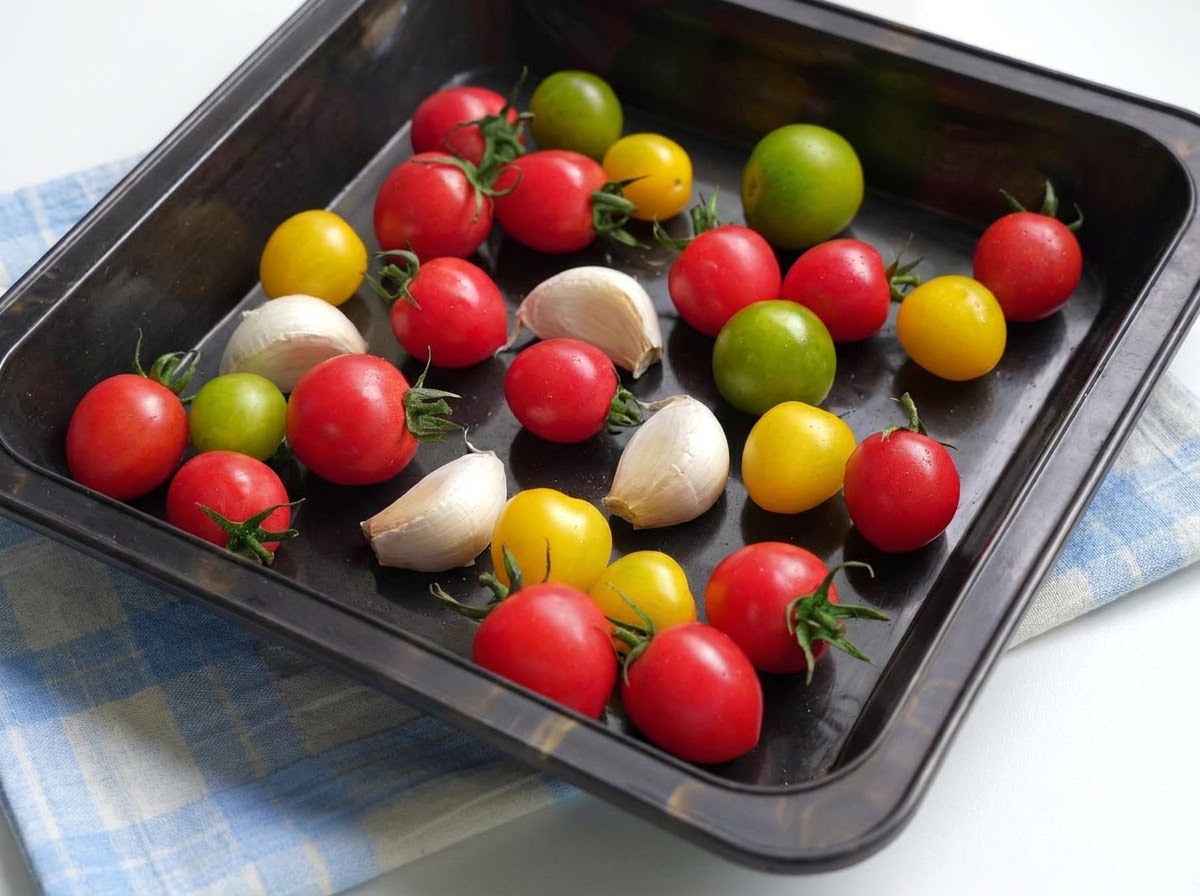 Une plaque de cuisson remplie de tomates cerises colorées et de gousses dail enrobées dhuile dolive et dassaisonnements.