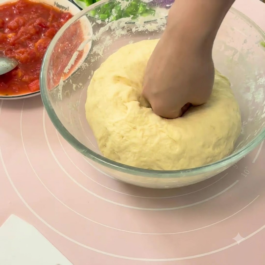 Une main enfonçant une grande boule de pâte à pizza levée à lintérieur dun bol en verre.