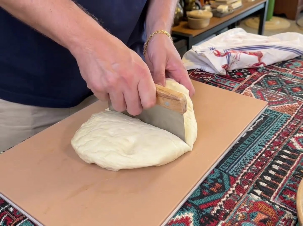 Un racloir à pâte coupant un grand monticule de pâte à pizza blanche en quatre morceaux séparés sur une planche à découper.