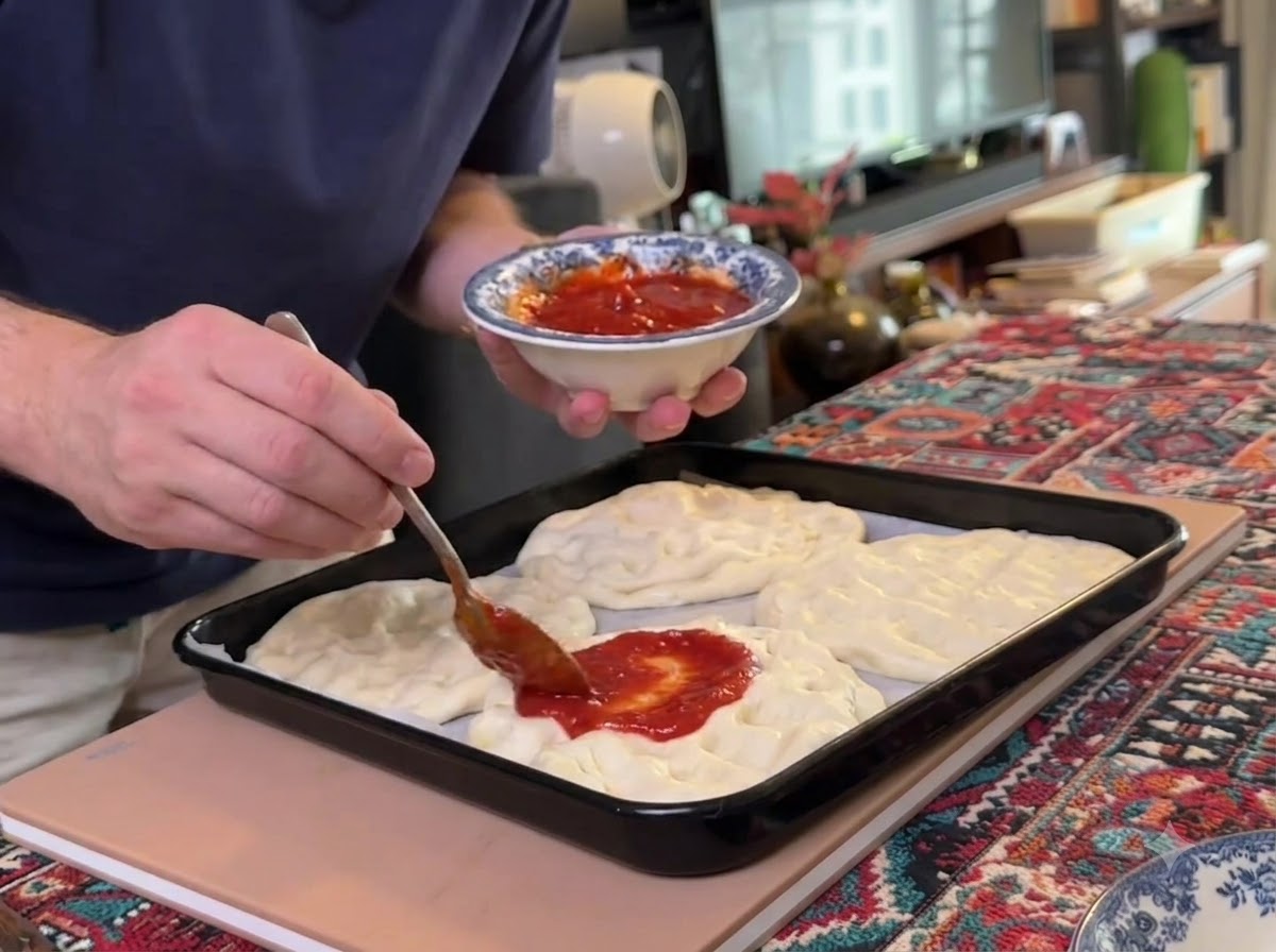 Une cuillère étalant de la sauce tomate rouge sur plusieurs bases de pâte à pizza aplaties disposées sur une plaque de cuisson noire.