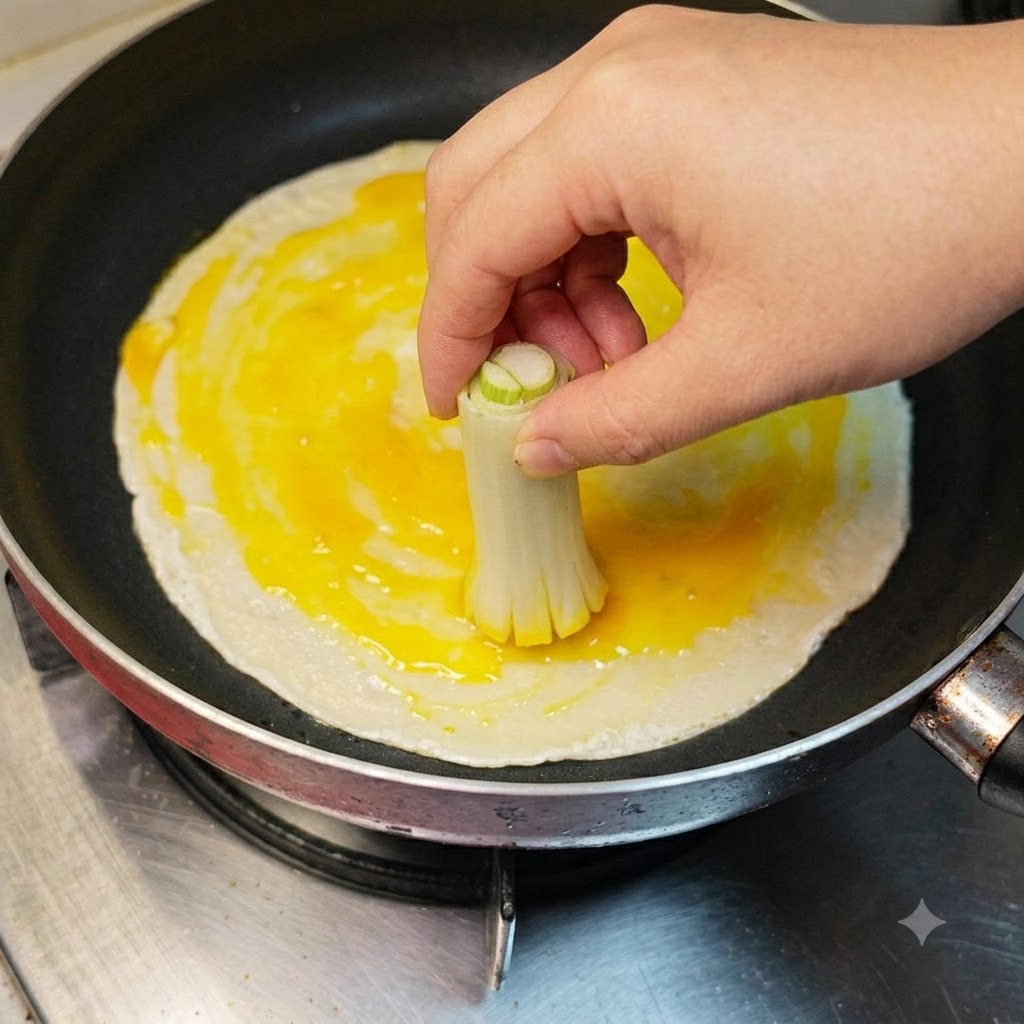 Une main utilisant un bouquet de tiges de ciboule attachées pour badigeonner le blanc et le jaune dœuf sur une crêpe.