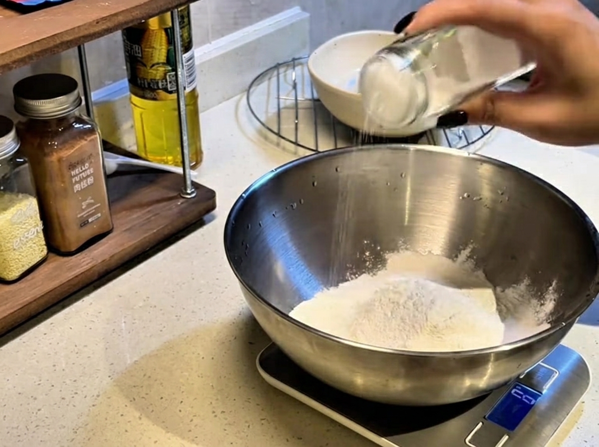 Une main secouant un bocal en verre pour saupoudrer du sel sur un dôme de farine blanche à lintérieur dun grand bol mélangeur en métal.