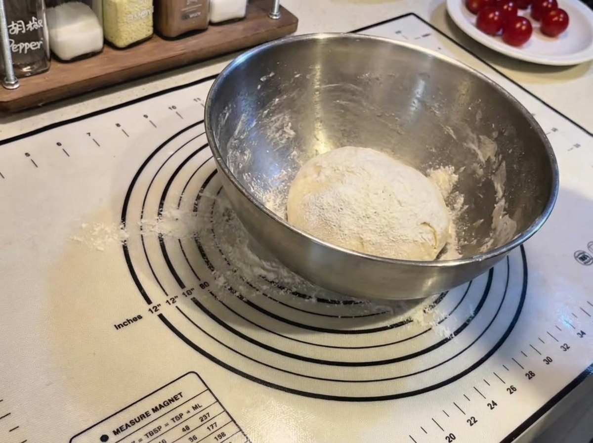Une boule de pâte à pizza ronde saupoudrée de farine à lintérieur dun bol en acier sur un tapis de cuisson en silicone.