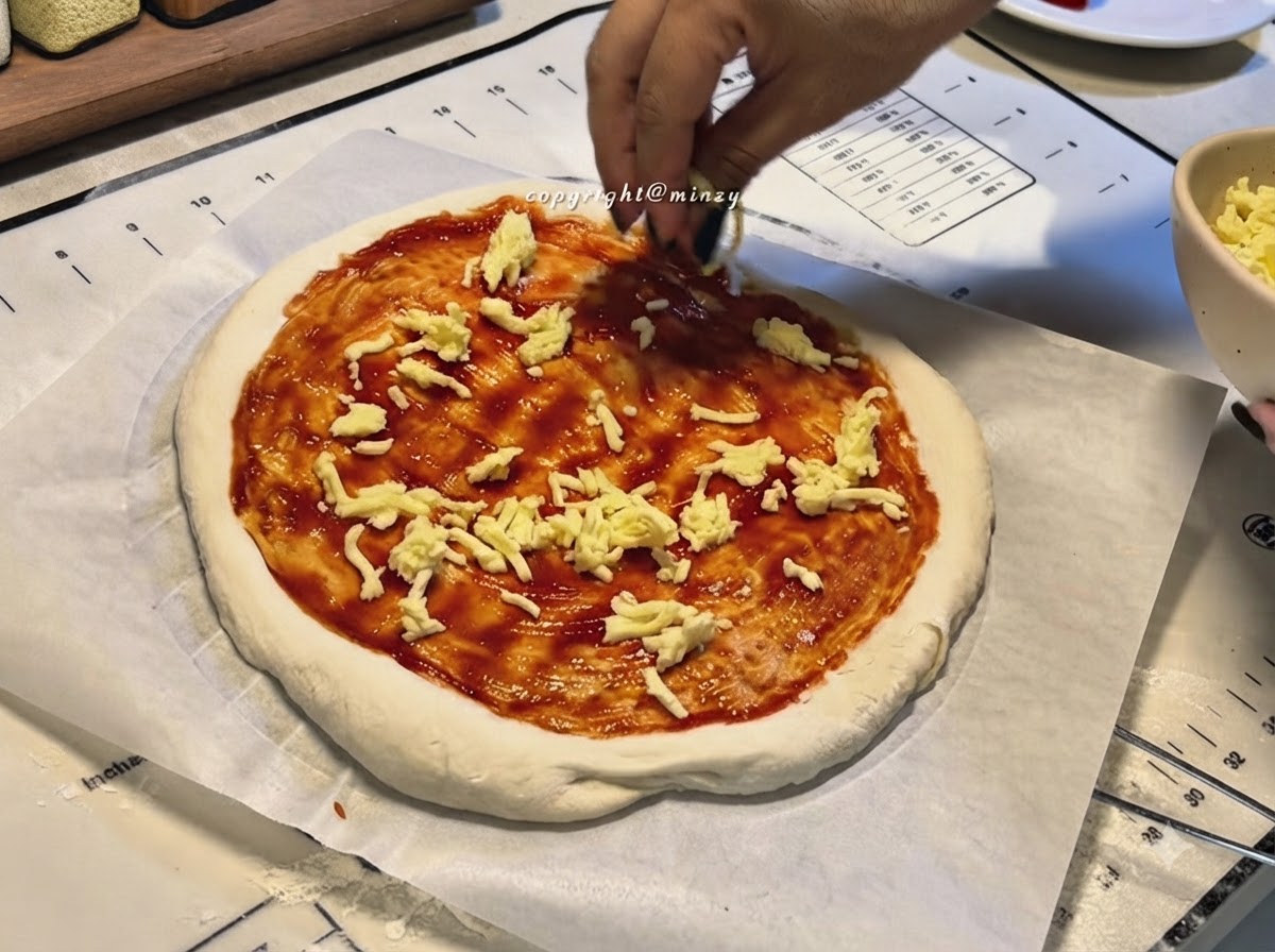 Des mains saupoudrant du fromage râpé sur une base de pizza recouverte de sauce tomate rouge.