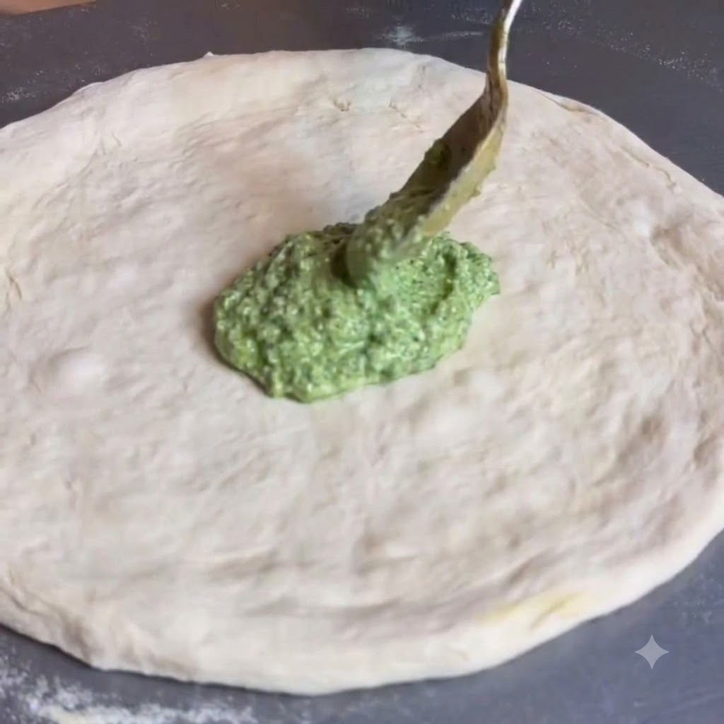 Une cuillère étalant de la sauce pesto vert au centre dune pâte à pizza ronde et plate.
