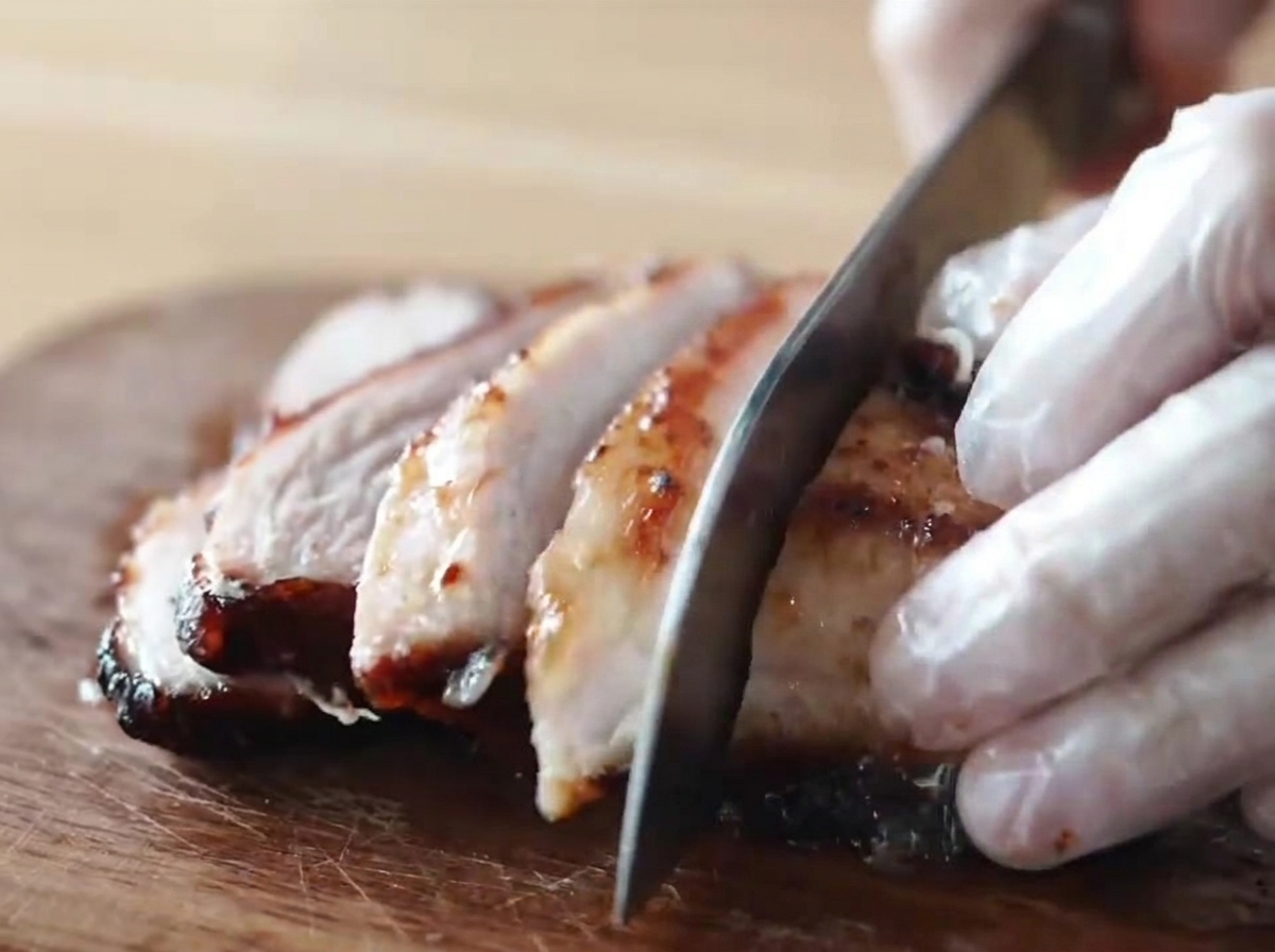Un morceau de porc chashu fumé laqué au miel et caramélisé, découpé en tranches épaisses par une personne portant des gants sur une planche à découper en bois.