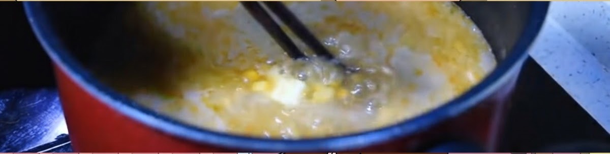 Des baguettes remuant un bouillon de miso orange en ébullition vigoureuse avec du maïs et une noisette de beurre dans une casserole sombre.